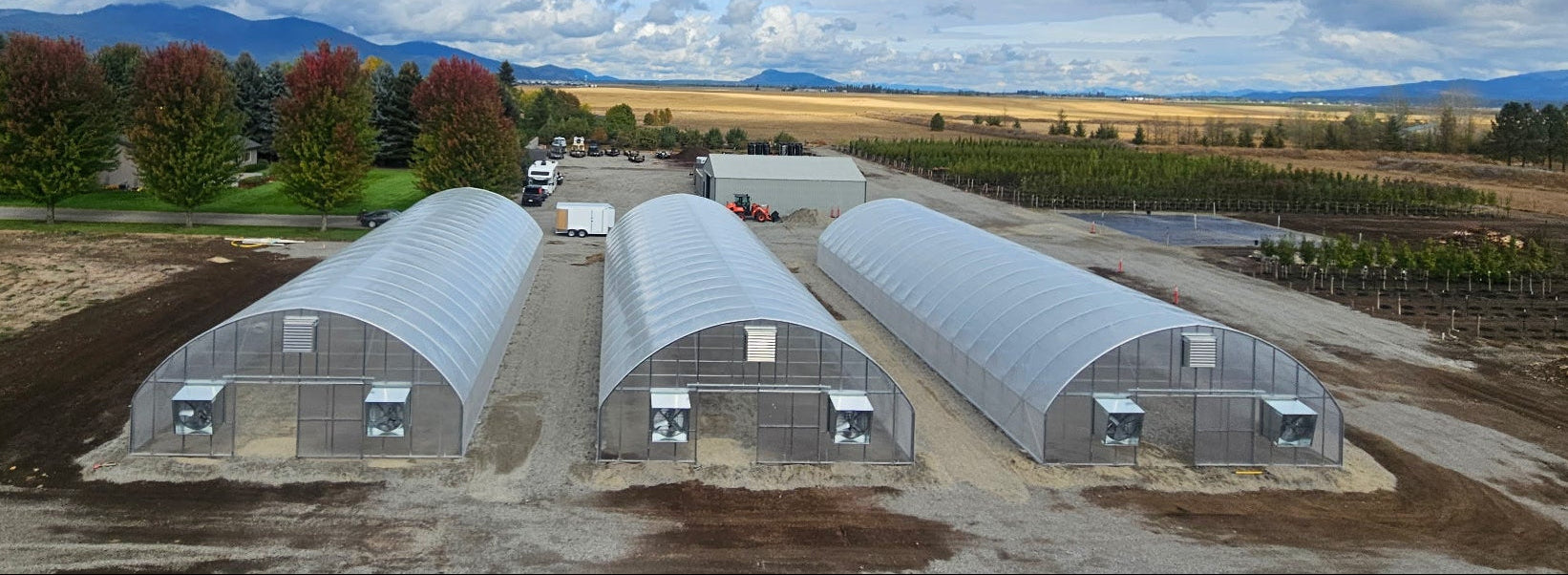 Three large greenhouses in a rural setting with farmland and mountains in the background.