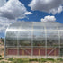 Clear greenhouse structure on a grassy area with a blue sky and clouds in the background.